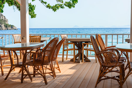 Rattan chairs and tables set on a seaside terrace overlooking clear blue water on a bright and sunny dayの写真素材
