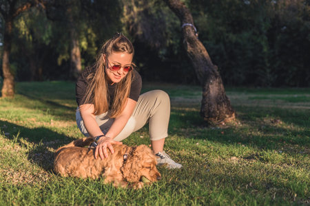 A young Caucasian woman with long brown hair and sunglasses interacts with a small, golden cocker...