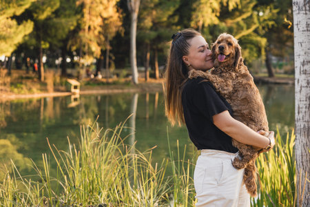 A young Caucasian woman with long brown hair holds a small brown cocker spaniel by a lake surrounded...