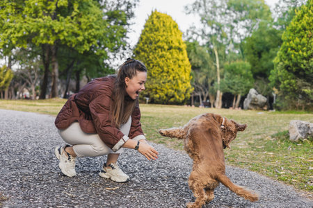 A young woman with long brown hair plays with a small brown dog in a green park. The dog is jumping excitedly on a gravel path.の写真素材