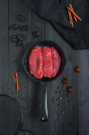 Raw fresh pieces of meat on the cast-iron pan, prune and oriental spices on the black wooden table の写真素材