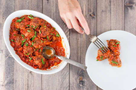 Female hand to take the peace of meatball with tomato sauce from the white plate, wooden table, top viewの写真素材