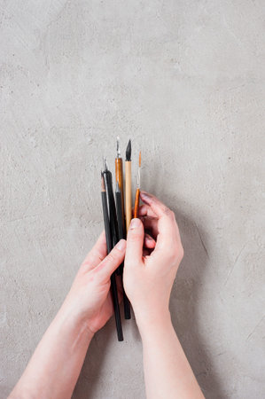 Woman's hands holding the art brushes and ink pens on the textured gray table, top view