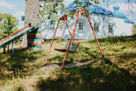 old metal colored swing for children on the street against the background of trees and churchの写真素材