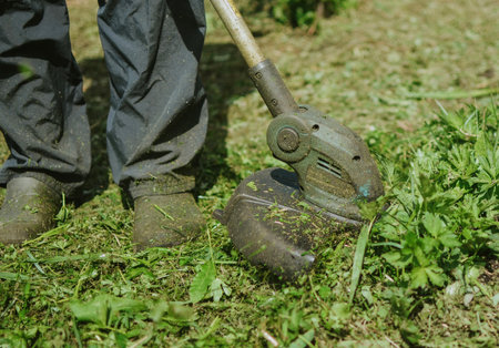 a man in pants and closed shoes mows overgrown grass with a lawn trimmer in the summer gardenの写真素材