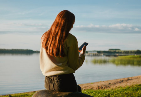 Young girl is sitting on a stone and watching her phone at the lake beach with clear summer waterの写真素材