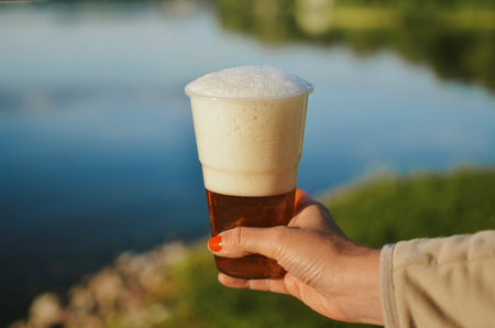holding by hand dark beer with foam against the background of a lake on a summer evening day