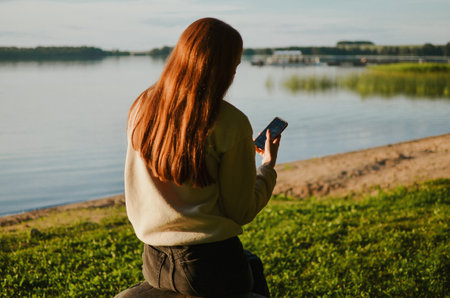 Young girl is sitting on a stone and holding her phone at the lake beach with clear summer waterの写真素材