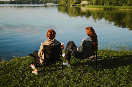 friends are sitting on camping chairs, enjoying a summer and drinking beer with foam by the lakeの写真素材