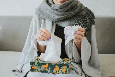 a woman wrapped in a scarf takes out tissues for her nose against the background of medicinesの写真素材