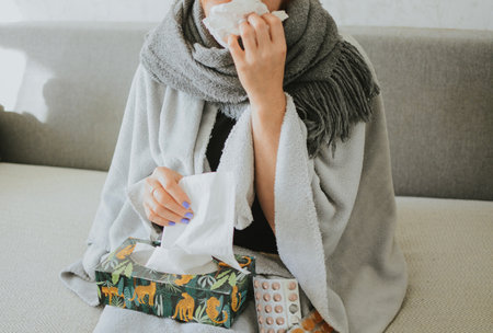 a woman wrapped in a scarf blows her nose with napkins against the background of medicinesの写真素材