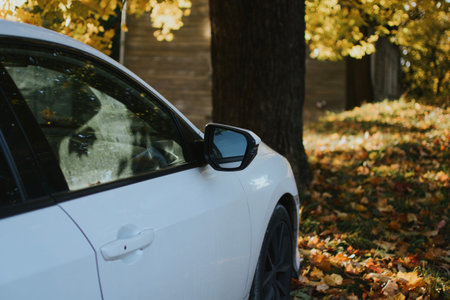 parked white car with side mirror near house with windows in park with fallen autumn yellow leavesの写真素材