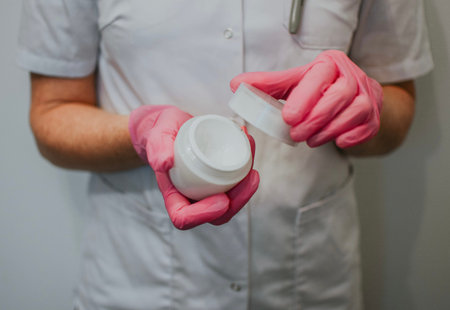A nurse in a white coat and pink gloves holds a white jar of cream for wounds and scratches on skin