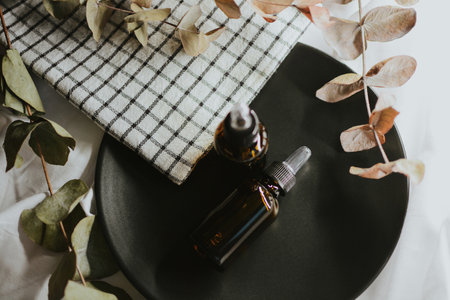 Brown cosmetic jars with pipette for serum close-up on a black plate with a checkered towel and eucalyptus on a white fabric background. High quality photoの写真素材