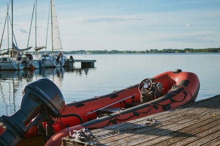 A red motorboat is moored to a wooden pier on a calm lake surface. The calm water reflects the sky and surrounding nature, creating an atmosphere of coziness and harmony. High quality photoの写真素材