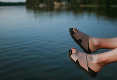 Women's feet in stylish sandals dangle from a wooden pier above the calm surface of the lake. A summer day filled with silence, relaxation, and enjoyment of the moment in nature. High quality photoの写真素材