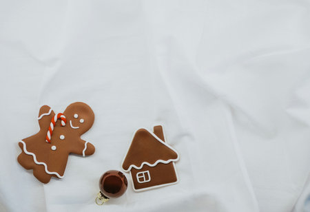 Gingerbread cookies in the shape of a gingerbread man and a house, decorated with icing, lie on white fabric next to a Christmas ornament. The composition creates a festive atmosphereの写真素材