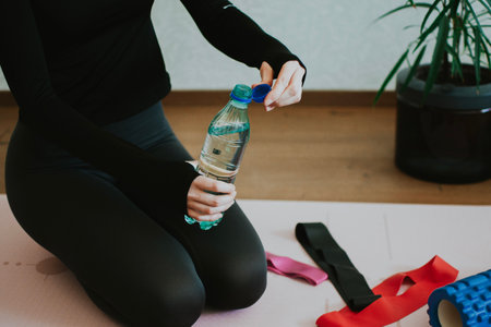 Woman sitting on exercise mat with resistance bands, foam roller for pilates at home and holding bottle of water to stay hydrated. High quality photoの写真素材