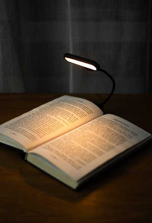 Reading a book at a wooden desk in the evening with the help of a convenient clip-on lamp. Eye...