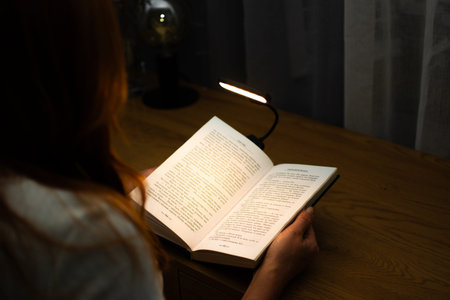 A woman is reading a book at a wooden table in the evening with the help of a convenient clip-on...