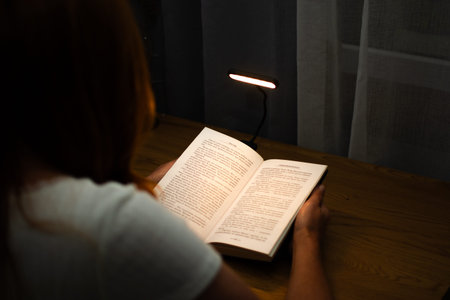 A woman is reading a book at a wooden table in the evening with the help of a convenient clip-on...