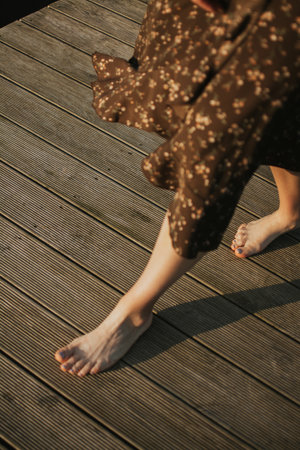 Barefoot woman walking on a wooden pier in summer Estonia, wearing a brown floral dress. Warm sunlight, natural atmosphere, and peaceful European nature. High quality photoの写真素材