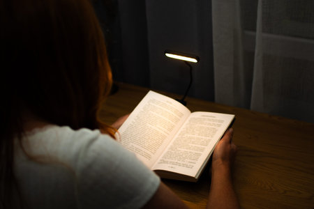 A woman is reading a book at a wooden table in the evening with the help of a convenient clip-on...