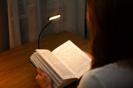 A woman is reading a book at a wooden table in the evening with the help of a convenient clip-on lamp. Eye protection in the dark and suitable warm light for health. High quality photoの写真素材