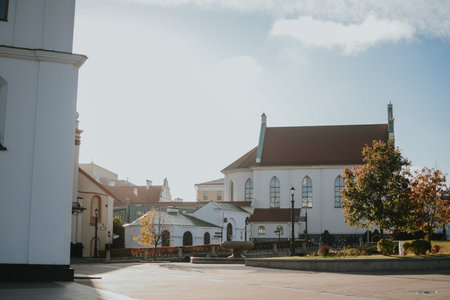White building with a green roof on the Town Hall Square in the Upper Town of Minsk. The morning autumn sun illuminates the facade and yellow trees, creating a warm city atmosphere. High quality photoの写真素材