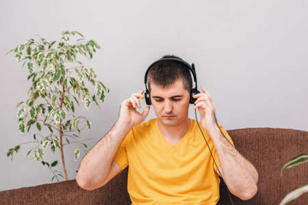 A young man in a yellow T-shirt talks on headphones, works from home and communicates with a client on the Internet. Room with houseplants. Concept of online education and homework, freelance.の写真素材