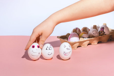 The child puts painted white eggs on a pink background. Painted faces by marker with different happy emotions. Minimal concept, and holiday and needlework. Copy space.の写真素材