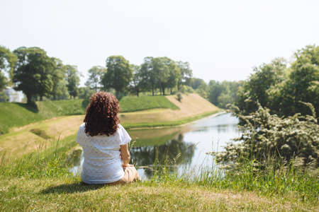 Girl Traveler sits on the green grass and looks at the lake. Rear view of a woman with curly hair who enjoys a beautiful view of the water. The concept of recreation in silence in natureの写真素材