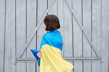 Young girl with blue and yellow state flag of Ukraine on her shoulders on a background of gray wooden wall on the street in snowy weather. Ukrainians against the war.の写真素材