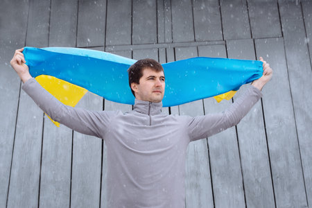 A man holds the state flag of Ukraine outdoors in snowy spring weather. Ukrainian flag, patriotism. Defender of the country, his land and family. Ukrainians against the war.の写真素材