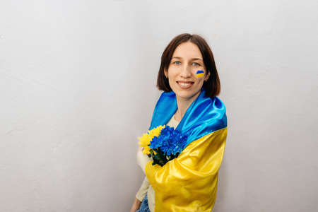 A young girl is wearing the national flag of Ukraine and holding flowers on a white background. The girl has a blue and yellow flag on her cheek. Independence Day of Ukraine. Copy Space.の写真素材