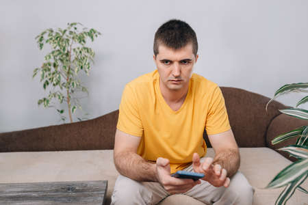 Young sad and angry man in a yellow T-shirt holding a phone in his hands. Green plants in the room.の写真素材