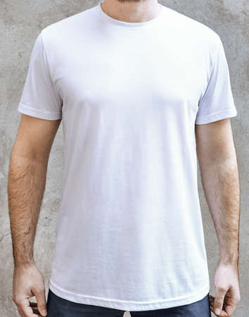 Young man in an empty white casual t-shirt. Front view close up on a background of light gray concrete wall. Design and layout of mens t-shirt for printing.の写真素材