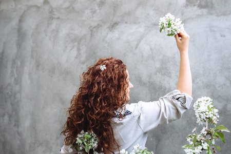 Beautiful girl with long curly brown hair in a linen shirt holds a flowering branch of cherries in...
