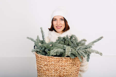 Young pretty girl smiling and holding lush Christmas tree branches in a wicker basket. Woman in white knitted sweater, hat and gloves on wall background. Minimalist Concept of Christmas and New Year.の写真素材