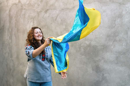 Soft selective focus on a smiling girl holding blue and yellow national flag of Ukraine. Woman in traditional embroidered shirt on a background of textured concrete wall. Independence Day. Copy spaceの写真素材