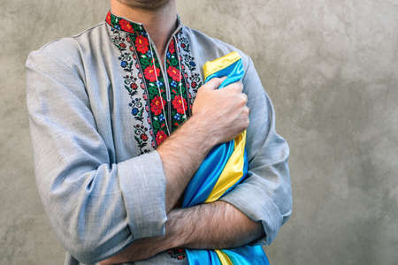 Soft Selective focus Man holds the blue and yellow national flag of Ukraine over his heart. Guy in a traditional embroidered shirt on a background of textured concrete wall. Copy spaceの写真素材