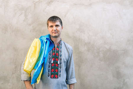 Portrait of a man holding the blue and yellow national flag of Ukraine on his shoulder and looking away. The guy in a traditional embroidered shirt on a background of a concrete wall. Copy space.の写真素材