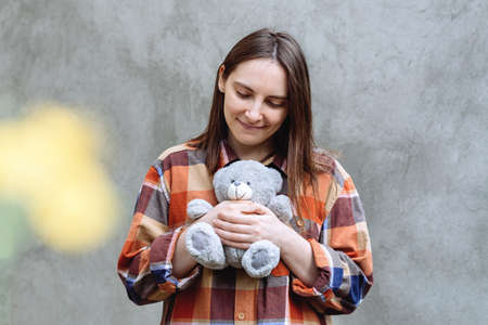 Girl holding a cute teddy bear toy in her hands. Woman in a plaid shirt on a background of gray concrete wall. Yellow spot on the wall. The brunette looked down.の写真素材