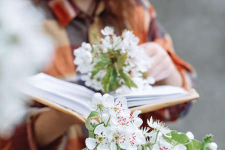 Soft selective focus apple blossoms bloom in the foreground. Girl holding a close up book wrapped in kraft paper on a gray background. Woman in a plaid shirt. Spring conceptの写真素材