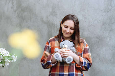 Girl holding a cute teddy bear toy. Woman in a checkered shirt on a background of gray concrete wall. Yellow spot and spring flowers on the wall. The brunette looked down.の写真素材