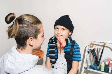 Children draw on their faces neat thin black French mustaches with a brush. Hat and scarf on the girl. An alternative image for Halloween. Real people. Have fun at home. Copy spaceの写真素材