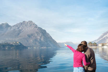 Soft focus, back view of couple enjoying a romantic date on the shores of Lake Lecco, Italy. The girl indicates direction with her hand. People in love on background of mountains, water. Copy spaceの写真素材