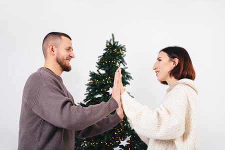 Young beautiful couple looking at each other on background of Christmas tree yellow lights. Lovers smile and hold hands. Woman in white knitted sweater. Minimalist concept of Christmas and New Year.の写真素材