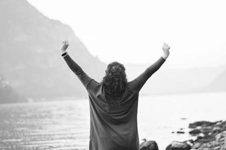 Black and white photo of happy girl standing on the beach and enjoying solitude on shores of Lake Lecco, Italy against the backdrop of mountains, water outdoors. Adventures. Blurred focus, rear viewの写真素材