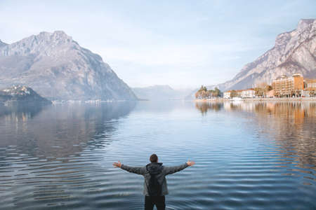 Blurred focus. Man traveler enjoys freedom with raised hands and backpack on the shores of Lake Lecco, Italy. Blue water, mountains and sky. Concept of tourist lifestyle, summer vacation outdoors.の写真素材